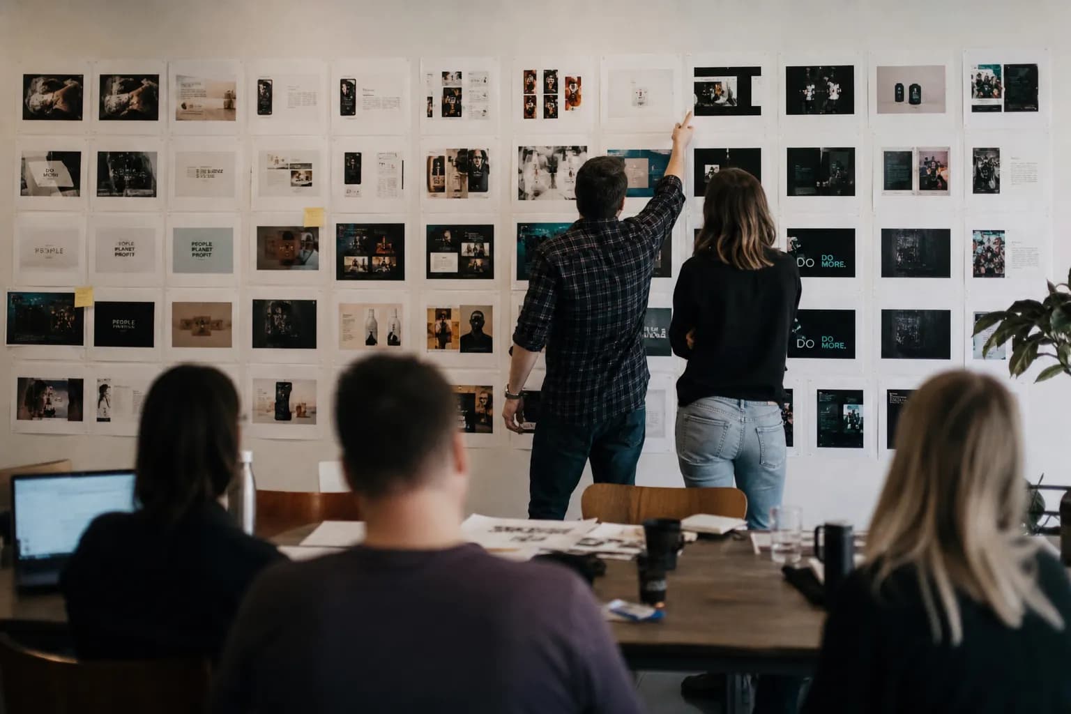 An agency team seen from behind reviewing a wall of printed ad mockups arranged in rows, two people standing at the wall pointing at one row