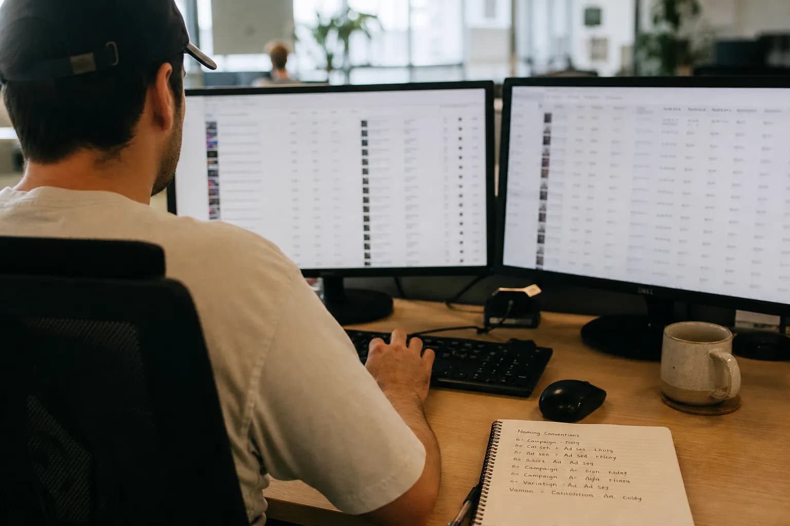 A performance marketer at a desk mid-launch in Ads Manager, multiple ad-set duplicates and creative variants open across two screens